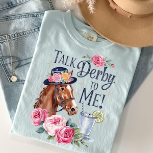 Może przedstawiać: Jasnoniebieska koszulka z grafiką konia w kapeluszu w kwiaty i napisem "Talk Derby to Me!". Projekt zawiera r&oacute;żowe r&oacute;że i koktajl. Beżowy kapelusz i niebieskie dżinsy w tle.