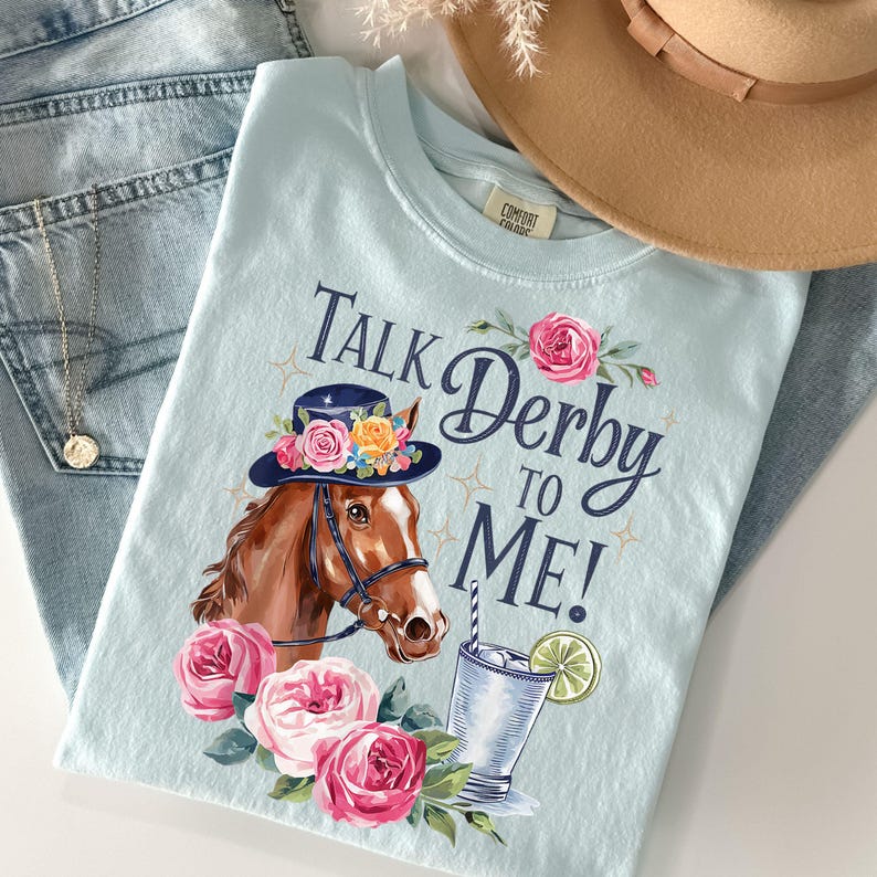 Może przedstawiać: Jasnoniebieska koszulka z grafiką konia w kapeluszu w kwiaty i napisem "Talk Derby to Me!". Projekt zawiera r&oacute;żowe r&oacute;że i koktajl. Beżowy kapelusz i niebieskie dżinsy w tle.
