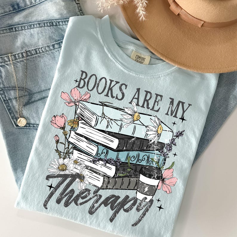 May include: A light blue t-shirt with the text "BOOKS ARE MY Therapy" and a graphic of stacked books, flowers, and a coffee cup. The shirt is laid on top of blue jeans, a necklace, and a tan hat.