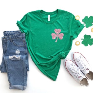 May include: A green t-shirt with a pink glitter shamrock design. The shirt is paired with blue jeans and white sneakers.