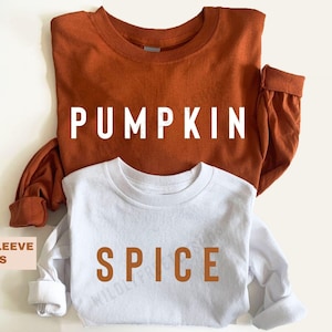 May include: A rust-coloured long-sleeve shirt with the word PUMPKIN printed in white letters and a white long-sleeve shirt with the word SPICE printed in rust-coloured letters. Both shirts feature a crew neckline and long sleeves with cuffs.