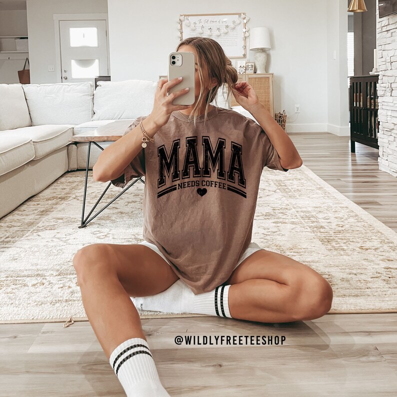 May include: A brown short-sleeved t-shirt featuring the black text MAMA NEEDS COFFEE with a small heart icon. The shirt has a crew neckline and a relaxed fit. The design is printed in a bold, collegiate-style font across the chest area.