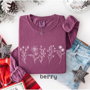 May include: Berry-colored long-sleeve sweatshirt with a white floral design across the front. The word "berry" is printed at the bottom. A silver necklace with a sun pendant is visible. The sweatshirt is paired with blue jeans.