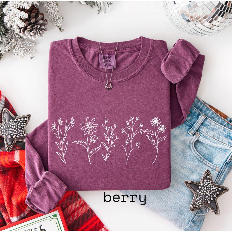 May include: Berry-colored long-sleeve sweatshirt with a white floral design across the front. The word "berry" is printed at the bottom. A silver necklace with a sun pendant is visible. The sweatshirt is paired with blue jeans.