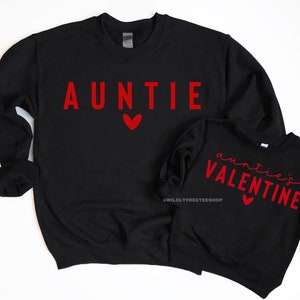 Może przedstawiać: Dwie czarne bluzy z czerwonym nadrukiem. Większa bluza ma napis "AUNTIE" z czerwonym sercem. Mniejsza bluza ma napis "auntie's VALENTINE" z czerwonym sercem.