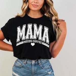 May include: A black short-sleeved t-shirt featuring white distressed lettering that reads MAMA NEEDS COFFEE with a small heart icon below the text. The design is centred on the chest and includes the text WILDLYFREETEESHOP in the upper right.