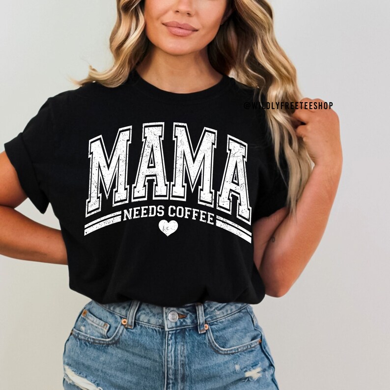 May include: A black short-sleeved t-shirt featuring white distressed lettering that reads MAMA NEEDS COFFEE with a small heart icon below the text. The design is centred on the chest and includes the text WILDLYFREETEESHOP in the upper right.