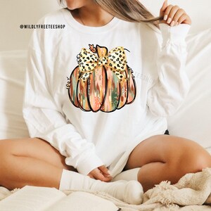 May include: White long-sleeve shirt featuring a colorful pumpkin design with a leopard print bow. The pumpkin has a black outline and is filled with orange, green, and pink brushstrokes. The text "@WILDLYFREETEESHOP" is visible in the upper left corner.