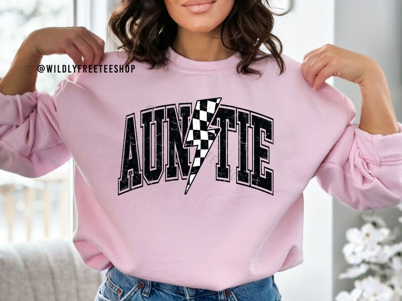 K&ouml;nnte beinhalten: Ein hellrosa Sweatshirt mit Rundhalsausschnitt und dem Wort AUNTIE in schwarzer College-Schrift mit abgenutzter Textur. Der Buchstabe I ist durch einen schwarz-wei&szlig; karierten Blitz ersetzt. Der Text @WILDLYFREETEESHOP erscheint in der Ecke.