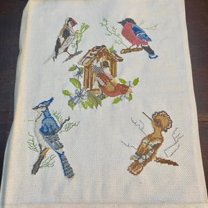 Op de afbeelding: Handgestikte stof met een kruissteekontwerp van verschillende vogels, een vogelhuisje en bloemmotieven. De vogels zijn afgebeeld in blauw, rood, bruin en grijs, zittend op takken tegen een crèmekleurige achtergrond.