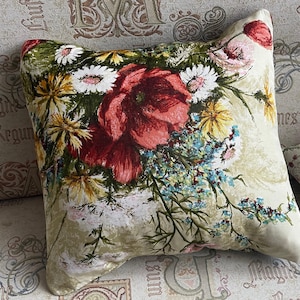 Può includere: Un cuscino decorativo con un motivo floreale con papaveri rossi, margherite gialle e fiori selvatici blu su uno sfondo beige.