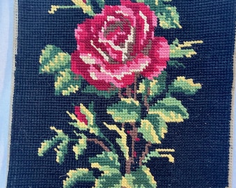 May include: A vintage needlepoint picture of a single pink rose with green leaves on a black background.
