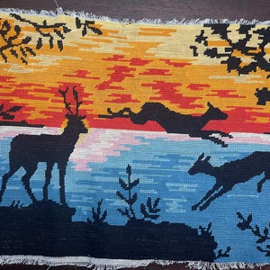 Op de afbeelding: Een tapijt dat een zonsondergangscène met herten afbeeldt. De lucht gaat over van geel naar rood, met zwarte silhouetten van bomen en herten. Eén hert staat, terwijl twee anderen over een blauwe en roze waterpartij springen. Het tapijt heeft een gefranjerde rand.