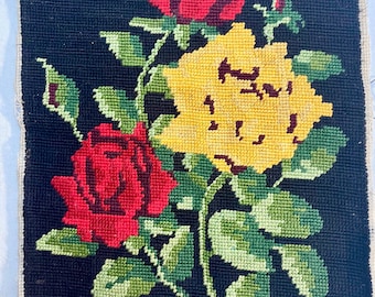 May include: A needlepoint floral design featuring two red roses and one yellow rose with green leaves and stems, set against a black background. The needlepoint is framed with a white border.