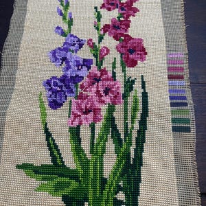May include: Needlepoint canvas featuring a floral design with purple and pink gladiolus flowers, green stems, and leaves. The flowers are stitched on a beige background. A color palette is visible on the right side.