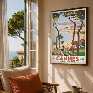 May include: A framed vintage travel poster for Cannes, France, featuring a coastal scene with palm trees and the text "Cannes Cote D'Azur". The poster is in a dark wood frame and hangs on a beige wall.