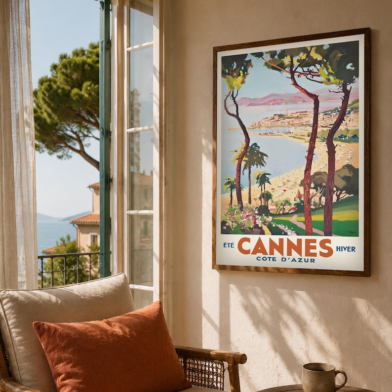 May include: A framed vintage travel poster for Cannes, France, featuring a coastal scene with palm trees and the text "Cannes Cote D'Azur". The poster is in a dark wood frame and hangs on a beige wall.