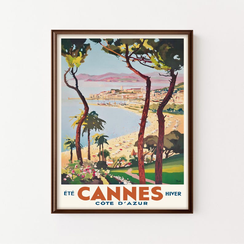 May include: A framed vintage travel poster featuring a coastal scene of Cannes, France. The artwork depicts a beach with people, palm trees, and buildings, with the text "&Eacute;T&Eacute; CANNES HIVER C&Ocirc;TE D'AZUR" in orange and white.