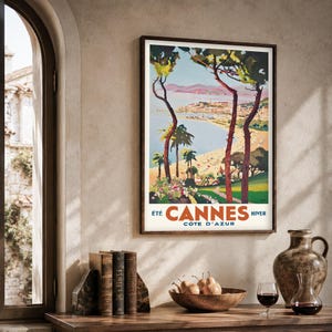 May include: A framed vintage travel poster for Cannes, France, featuring a beach scene with palm trees and the text "Cannes Cote D'Azur". The poster is in a dark wood frame, displayed on a wooden table with books, a bowl of pears, and a decorative vase.