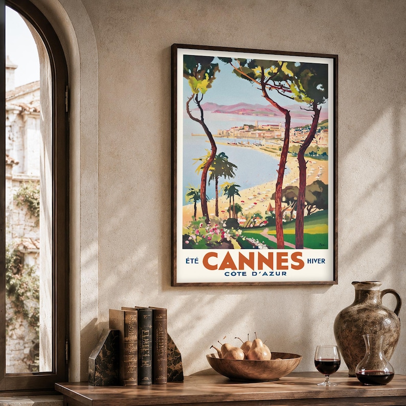 May include: A framed vintage travel poster for Cannes, France, featuring a beach scene with palm trees and the text "Cannes Cote D'Azur". The poster is in a dark wood frame, displayed on a wooden table with books, a bowl of pears, and a decorative vase.