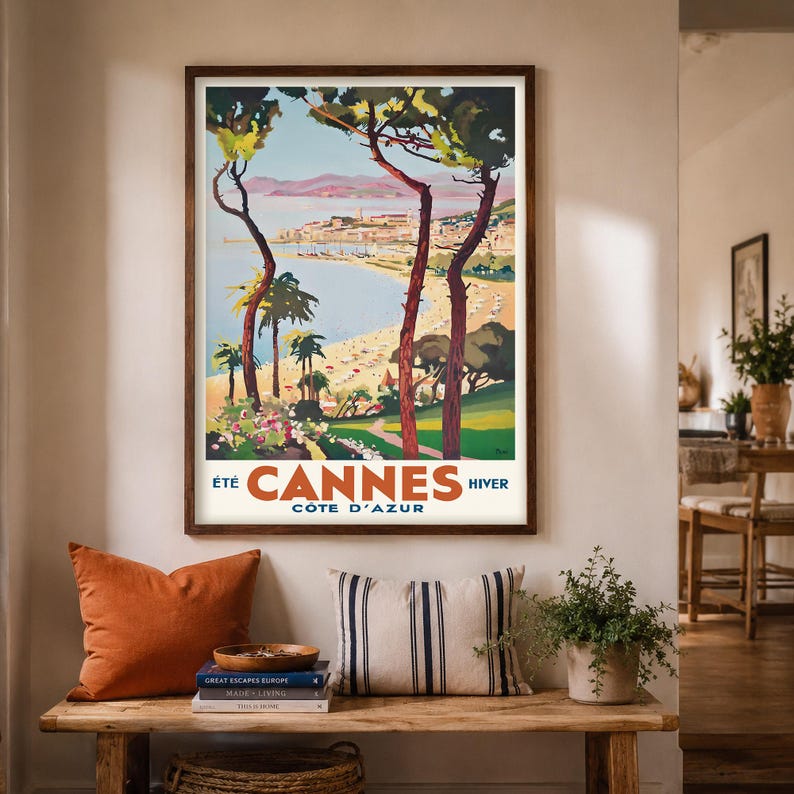 May include: A framed vintage travel poster for Cannes, France, featuring a beach scene with palm trees and the text "Cannes Cote D'Azur". The poster is in a dark wooden frame and hangs above a wooden bench with pillows and books.