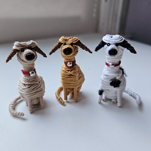 May include: Three wire-wrapped dog figurines. The dog on the left is beige, the dog in the middle is brown, and the dog on the right is white with black spots. Each dog has a red collar with a silver bell.