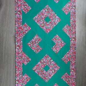 Può includere: Una trapunta rettangolare con un design geometrico verde e un bordo floreale. La trapunta presenta un motivo centrale di forme a diamante interconnesse in verde, con un bordo e accenti di piccoli fiori rosa e rossi.