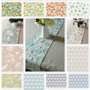 May include: A collection of 18 table runner designs featuring various patterns, including floral, geometric, and nature-inspired motifs. The table runners are displayed on a light grey linen tablecloth.