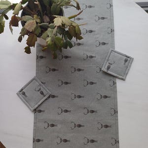 May include: A gray table runner with a repeating pattern of brown deer antlers. The runner is on a white table with a green plant in a white pot. Two gray square coasters with a deer antler design are on the table.