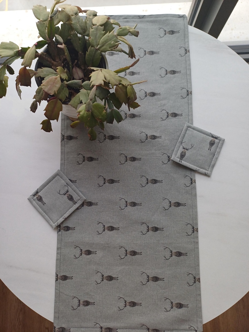 May include: A gray table runner with a repeating pattern of brown deer antlers. The runner is on a white table with a green plant in a white pot. Two gray square coasters with a deer antler design are on the table.