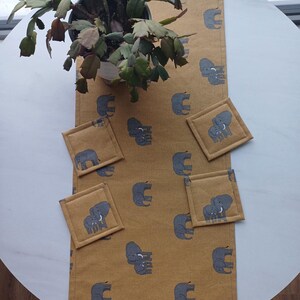 May include: A mustard yellow table runner with a repeating pattern of gray elephants. Four matching gray elephant coasters are placed on the runner.