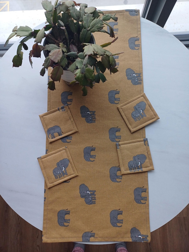 May include: A mustard yellow table runner with a repeating pattern of gray elephants. Four matching gray elephant coasters are placed on the runner.