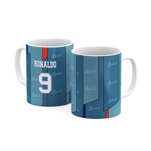 May include: A white ceramic mug with a blue and white design featuring the name "Ronaldo" and the number "9". The mug has a blue and white pattern with the word "Barca" repeated throughout.