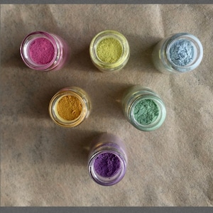 May include: Six small glass jars with lids containing different coloured powders. The powders are pink, yellow, orange, green, blue, and purple.