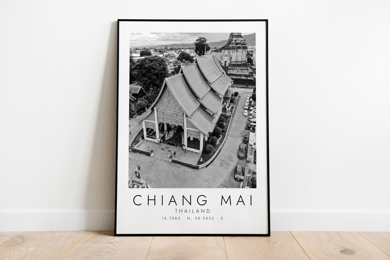 May include: Black and white photograph of a temple in Chiang Mai, Thailand. The temple has a traditional Thai roof and is surrounded by trees and a parking lot. The text "CHIANG MAI" is printed below the image, along with "THAILAND" and the coordinates "18.7883 N, 98.9833 E".