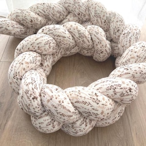 Könnte beinhalten: Ein geflochtener Baby-Bumper in runder Form. Der Bumper besteht aus cremefarbenem Stoff mit einem zarten Blumenmuster in Braun- und Beigetönen. Das Design ähnelt einem großen, weichen Kranz.