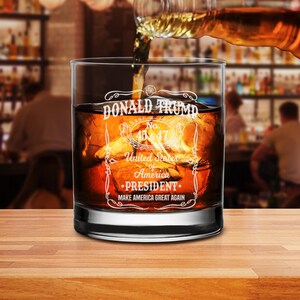 Puede incluir: Un vaso de whisky de vidrio transparente con el texto "DONALD TRUMP No. 45 & 47 United States of America PRESIDENT MAKE AMERICA GREAT AGAIN" grabado en el lateral. El vaso est&aacute; lleno de l&iacute;quido &aacute;mbar y cubitos de hielo.