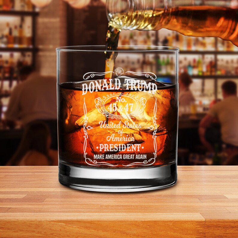 Puede incluir: Un vaso de whisky de vidrio transparente con el texto "DONALD TRUMP No. 45 & 47 United States of America PRESIDENT MAKE AMERICA GREAT AGAIN" grabado en el lateral. El vaso est&aacute; lleno de l&iacute;quido &aacute;mbar y cubitos de hielo.