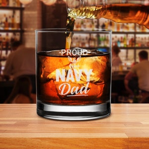 May include: A clear glass with a white inscription that reads "Proud Navy Dad" with a small Navy emblem. The glass is filled with brown liquid and ice cubes.