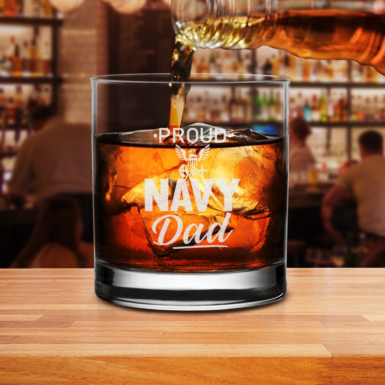 May include: A clear glass with a white inscription that reads "Proud Navy Dad" with a small Navy emblem. The glass is filled with brown liquid and ice cubes.