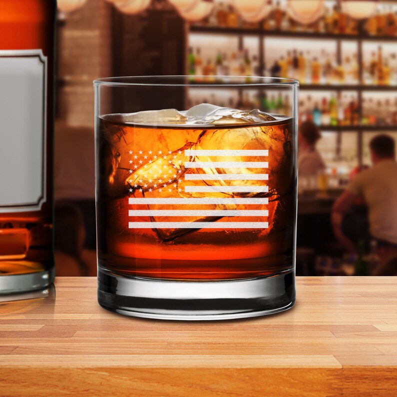 Puede incluir: Un vaso de rocas de vidrio transparente con un dise&ntilde;o de la bandera estadounidense grabado en el lateral. El vaso est&aacute; lleno de l&iacute;quido &aacute;mbar y cubitos de hielo.