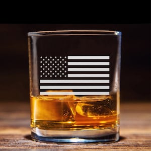Puede incluir: Un vaso de vidrio transparente con un dise&ntilde;o de bandera estadounidense grabado. El vaso est&aacute; lleno de l&iacute;quido &aacute;mbar y cubitos de hielo.
