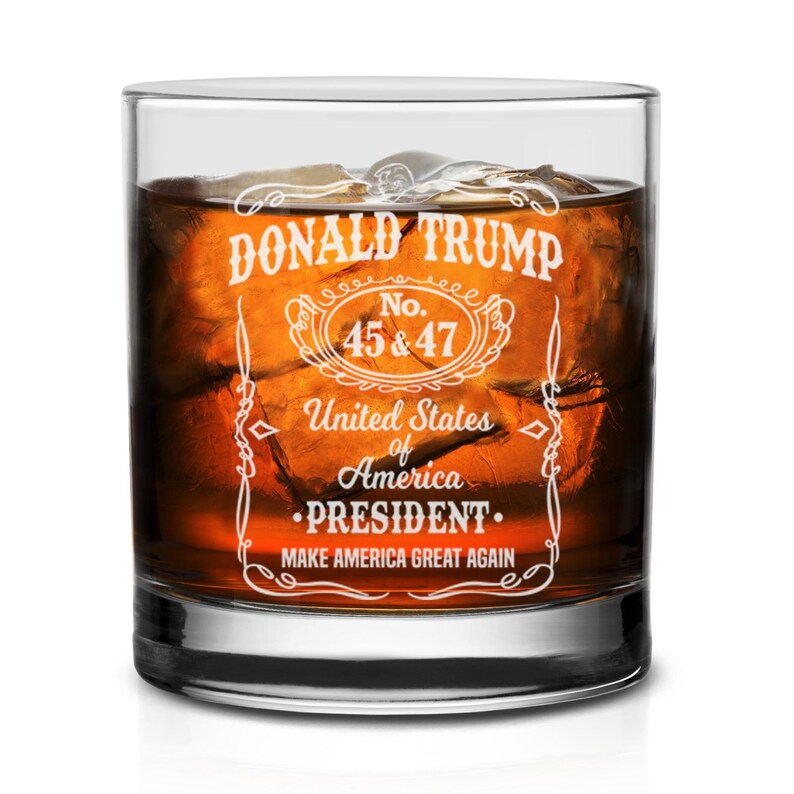 Puede incluir: Un vaso de vidrio transparente con un dise&ntilde;o blanco que dice "DONALD TRUMP No. 45 & 47 United States of America &bull;PRESIDENT. MAKE AMERICA GREAT AGAIN". El vaso est&aacute; lleno de un l&iacute;quido marr&oacute;n y cubitos de hielo.