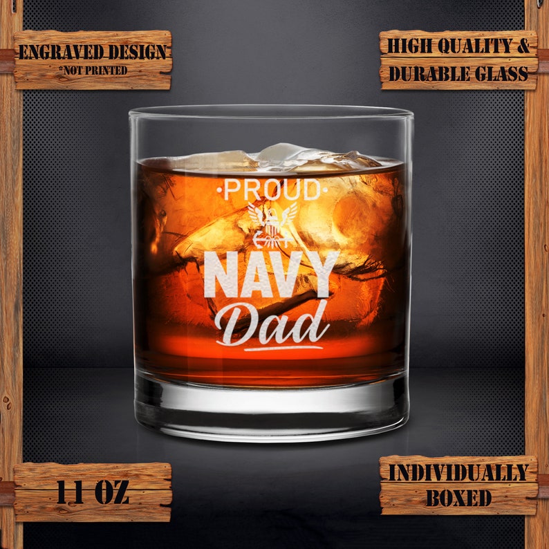 May include: An 11 ounce glass with an engraved design that says "Proud Navy Dad". The glass is filled with a brown liquid and ice cubes. The glass is on a dark background.