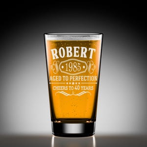 May include: A clear glass beer mug with a white inscription that reads "Robert 1985 Aged to Perfection Cheers to 40 Years".