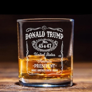 May include: A clear glass shot glass with a white design that says "DONALD TRUMP No. 45 & 47 United States of America •PRESIDENT• MAKE AMERICA GREAT AGAIN". The glass is filled with amber liquid.