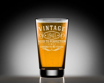 May include: A clear glass beer mug with a vintage design. The mug is filled with a golden yellow liquid and has the text "Vintage 1945 Aged to Perfection Cheers to 80 Years" etched on the side.
