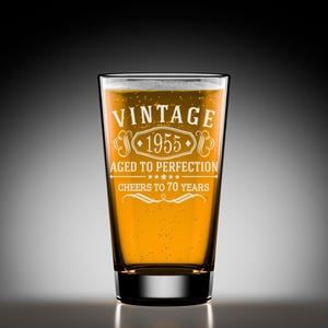 May include: A clear glass beer mug with a vintage design. The mug is filled with a golden yellow liquid and has the text "Vintage 1955 Aged to Perfection Cheers to 70 Years" etched on the side.
