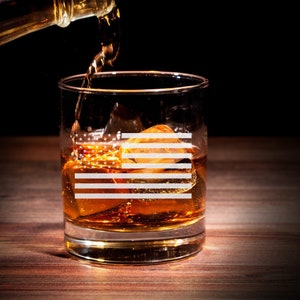 Puede incluir: Un vaso de vidrio transparente con un dise&ntilde;o de bandera estadounidense est&aacute; lleno de l&iacute;quido &aacute;mbar y cubitos de hielo. El vaso est&aacute; sobre una superficie de madera y una botella de l&iacute;quido &aacute;mbar se vierte en el vaso.