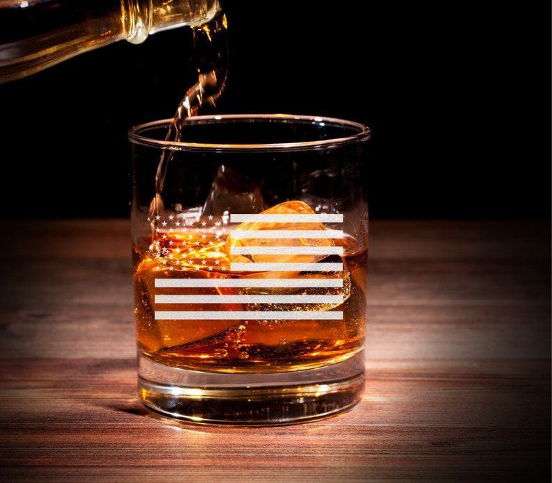 Puede incluir: Un vaso de vidrio transparente con un dise&ntilde;o de bandera estadounidense est&aacute; lleno de l&iacute;quido &aacute;mbar y cubitos de hielo. El vaso est&aacute; sobre una superficie de madera y una botella de l&iacute;quido &aacute;mbar se vierte en el vaso.